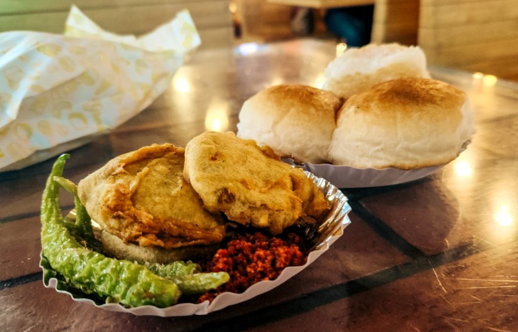 The image shows a classic vada pav—a deep-fried spiced potato fritter (vada) tucked inside a soft pav bun, served with green chutney and dry garlic chutney. Widely considered Mumbai’s most beloved street food, vada pav is affordable, flavorful, and quick to eat on the go. Often sold at roadside stalls across Maharashtra, it’s a vegetarian snack with bold flavors and a cult following. The photo captures the crispy texture and vibrant accompaniments that define this iconic Indian dish.