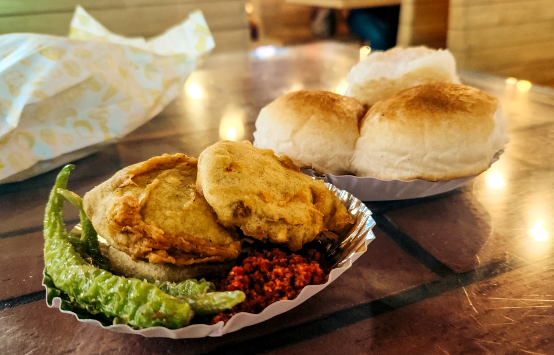 The image shows a classic vada pav—a deep-fried spiced potato fritter (vada) tucked inside a soft pav bun, served with green chutney and dry garlic chutney. Widely considered Mumbai’s most beloved street food, vada pav is affordable, flavorful, and quick to eat on the go. Often sold at roadside stalls across Maharashtra, it’s a vegetarian snack with bold flavors and a cult following. The photo captures the crispy texture and vibrant accompaniments that define this iconic Indian dish.
